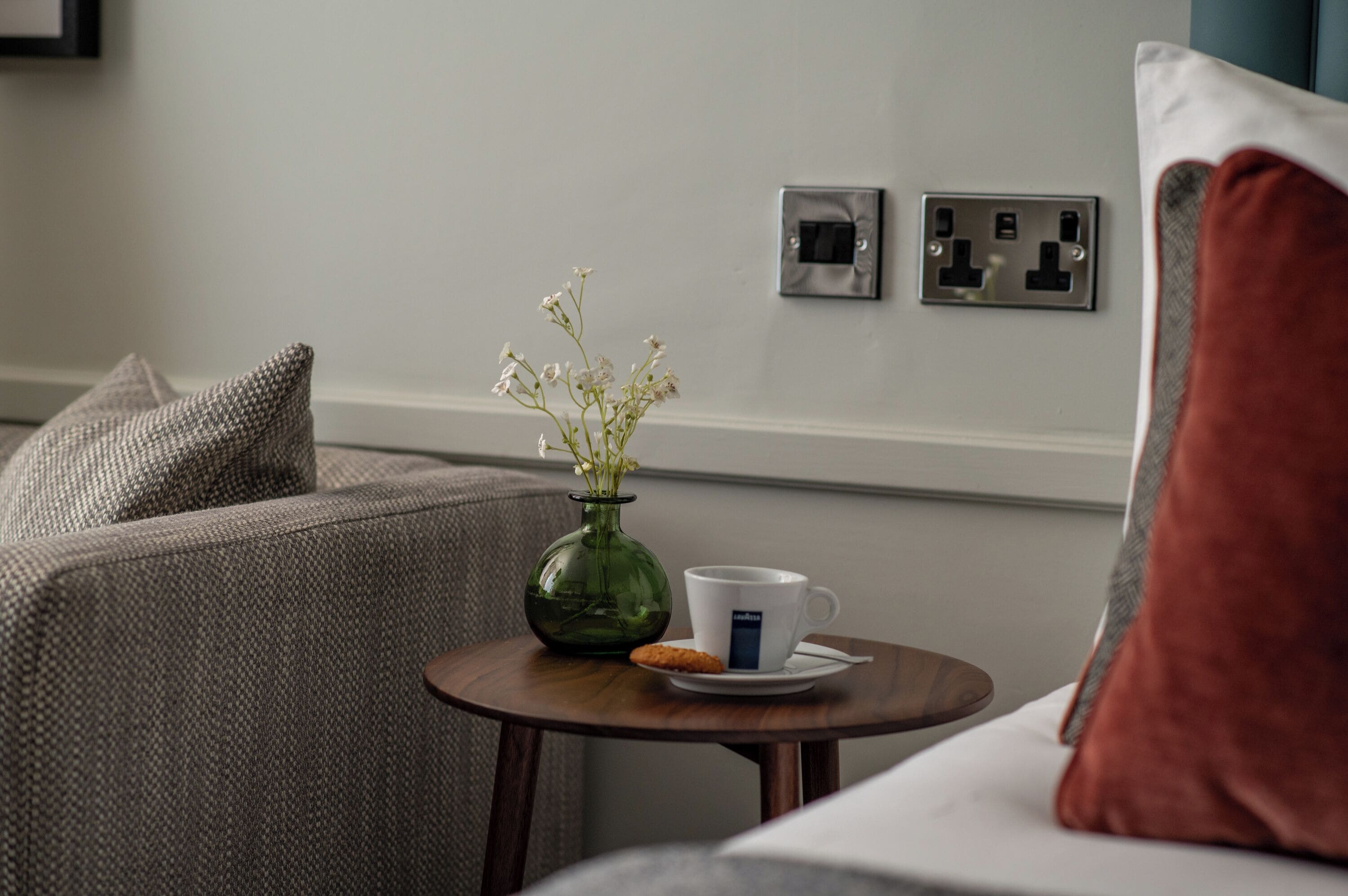 Une pièce avec un canapé gris, une petite table ronde sur laquelle se trouve un vase vert et une tasse blanche, et un coussin rouge.