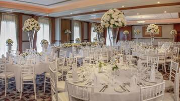 Une salle de banquet avec des tables rondes dressées pour un événement formel, agrémentée de chaises blanches et de centres de table floraux.