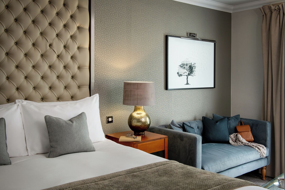 A hotel room with a tufted headboard, a bed with white linens, a grey sofa, and a framed picture on the wall.