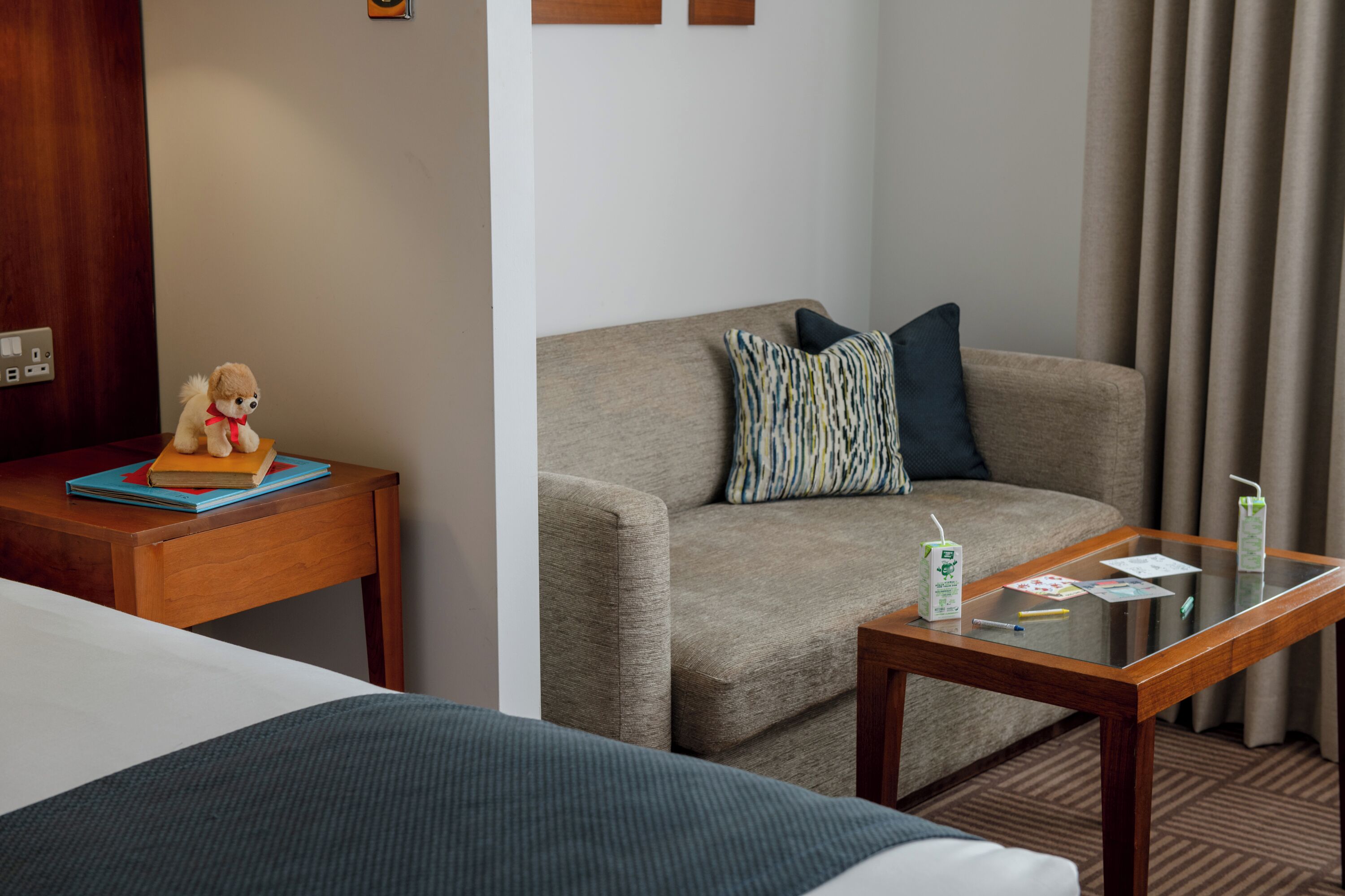 A hotel room with a bed, a wooden nightstand with a teddy bear and a book, a glass coffee table with a drink and cards, a beige sofa with striped pillows, and a window with curtains.
