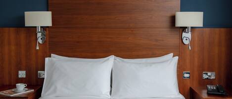 Habitación de hotel con cabecera de madera, dos lámparas de noche, cama blanca con dos almohadas, mesita de noche con una taza y un teléfono, y una manta oscura.