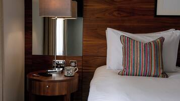 A hotel room with a wooden headboard, a bedside table with a lamp, a coffee maker, and a mug with the text "THAT'S I'LL DO!".