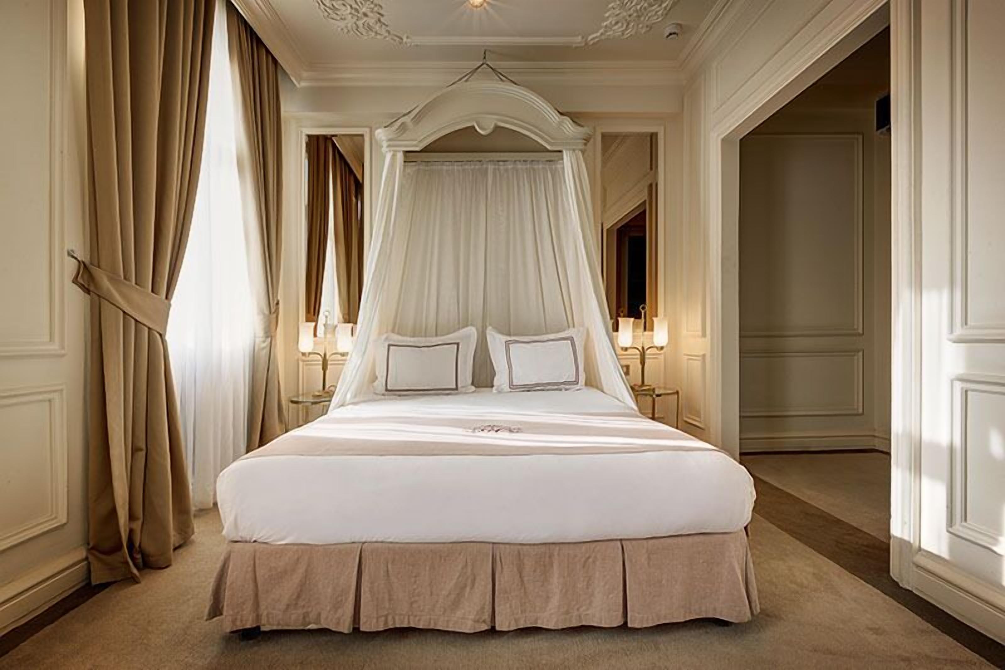 Una cama con dosel grande y ornamentada, ubicada en una habitación amplia con paredes color crema y piso alfombrado.