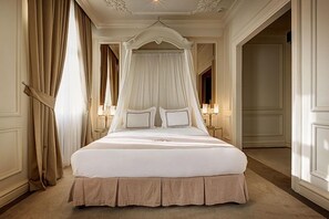 A large, ornate four-poster bed with a canopy, positioned in a spacious room with cream-colored walls and a carpeted floor.