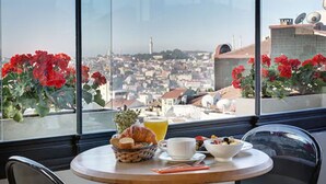 Une table de petit-déjeuner avec des croissants, des fruits et un bol de céréales, donnant sur un paysage urbain parsemé de fleurs rouges et offrant une vue sur les bâtiments.