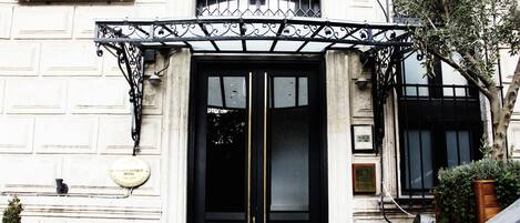 A hotel entrance with a black double door, a sign above the door, and a small garden area with potted plants.