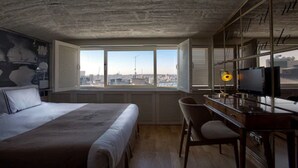 Une chambre d’hôtel avec un grand lit, un bureau avec une chaise, une télévision et une vue sur la ville grâce à de grandes fenêtres.