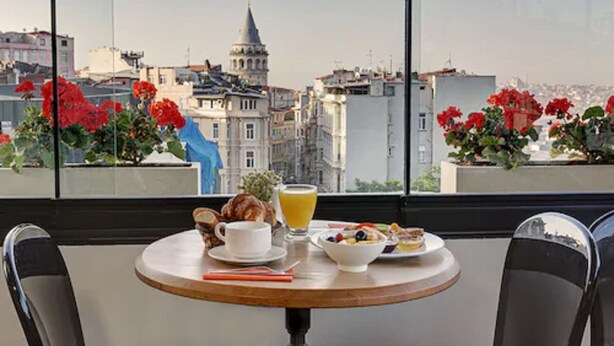 Une table avec des aliments pour le petit-déjeuner et un verre de jus d’orange, donnant sur un paysage urbain avec des bâtiments et des fleurs rouges.