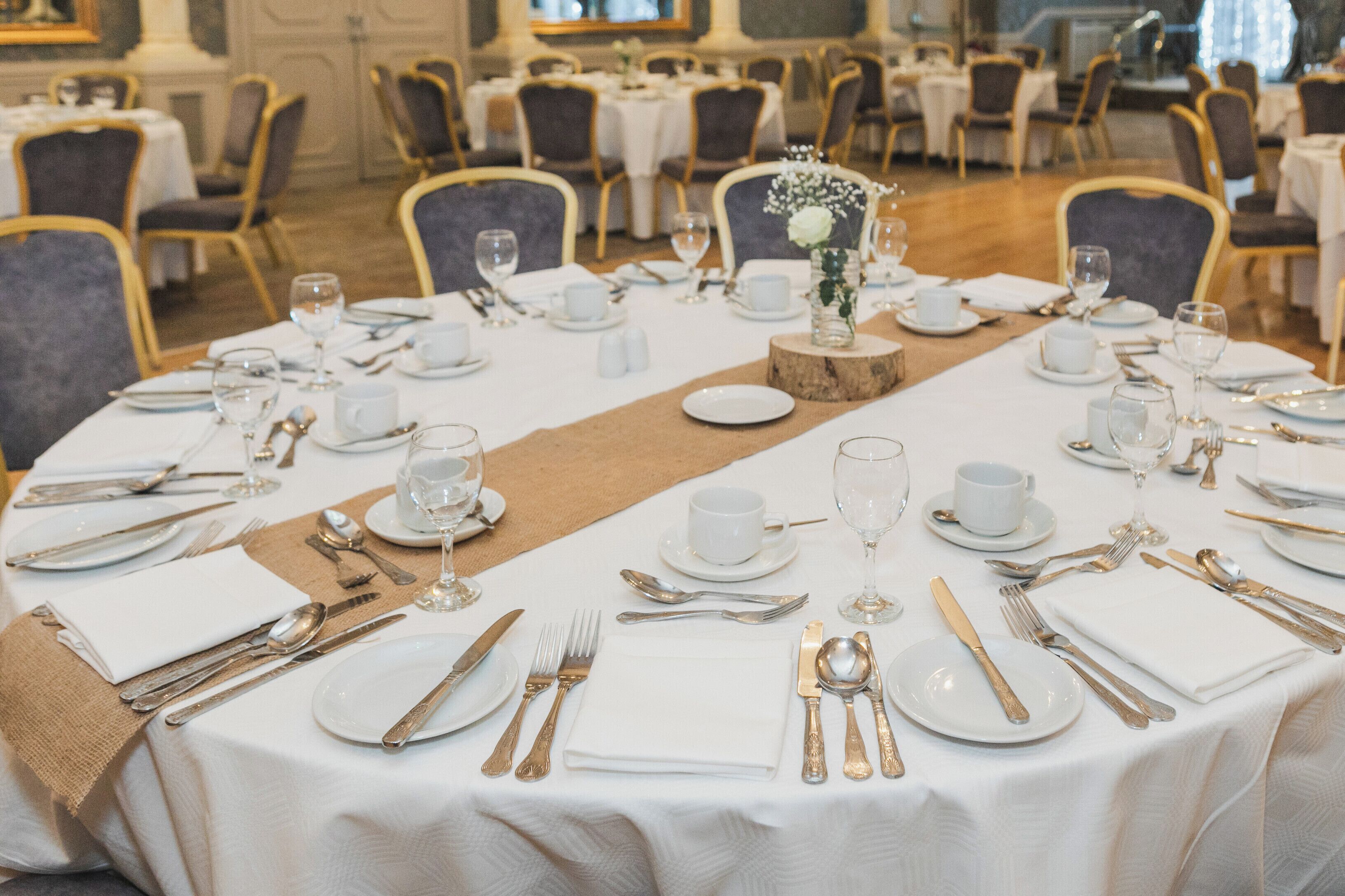 A banquet hall with round tables set for a formal event, featuring white tablecloths, gold cutlery, and floral centerpieces.