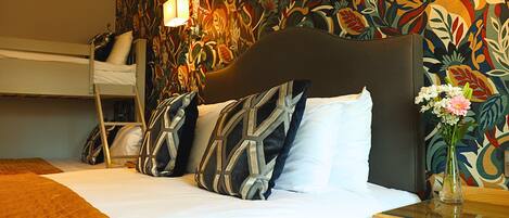 Une chambre d’hôtel avec un lit superposé, un grand lit agrémenté d’oreillers à motifs, une table de chevet avec un vase de fleurs et un mur recouvert d’un papier peint à motifs floraux.