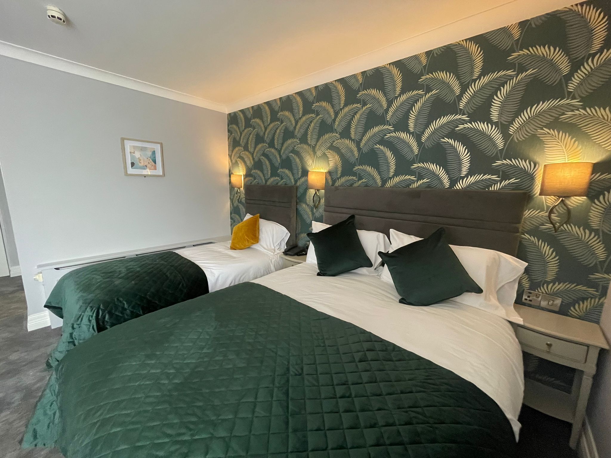 Ett hotellrum med två sängar, var och en med ett grönt, vadderat sängöverkast och vita sängkläder. Rummet har en stor vägg med en tapet med ett lövmönster och två vägglampor.