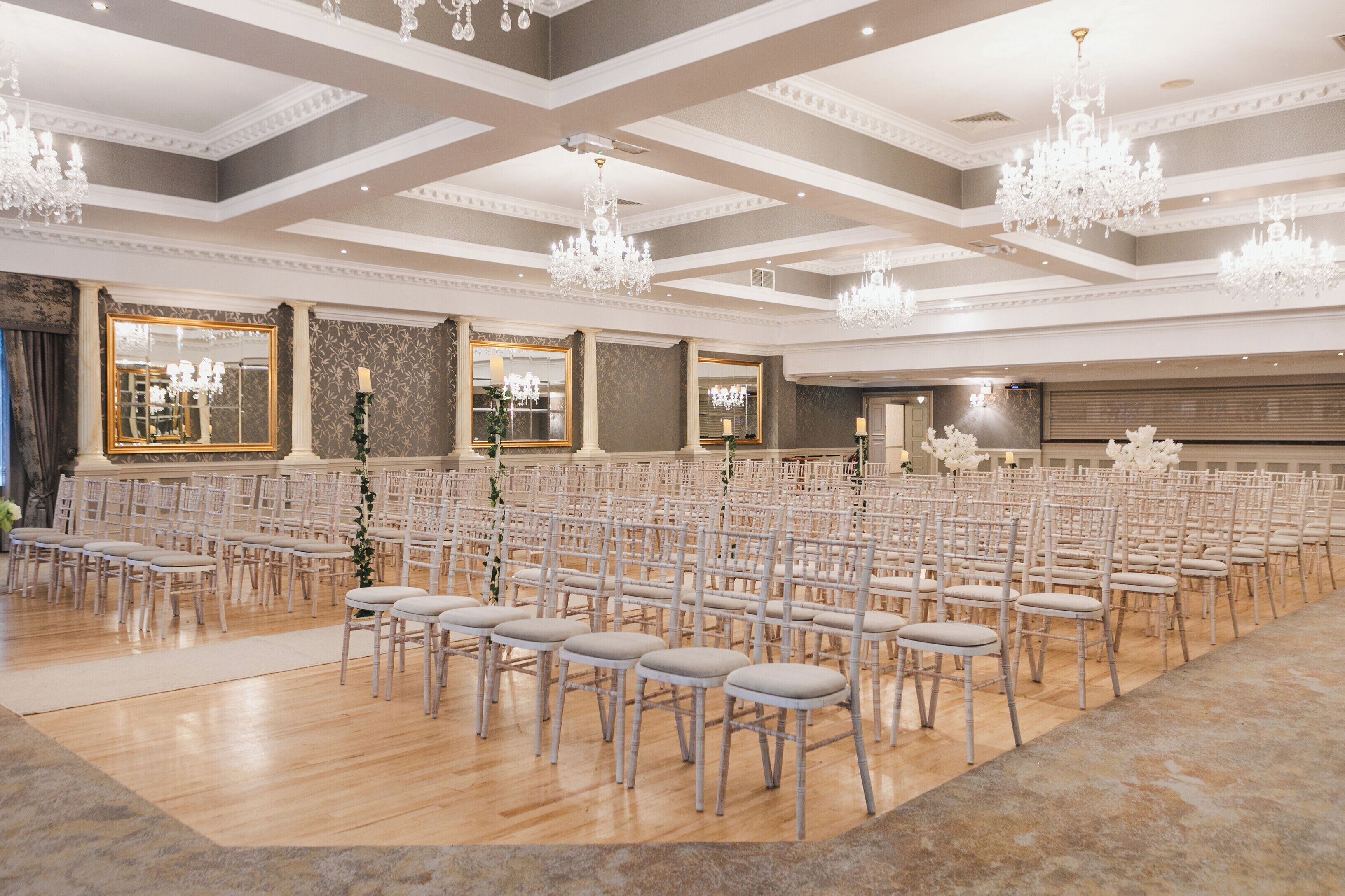 Un amplio salón con filas de sillas blancas, candelabros y espejos grandes.
