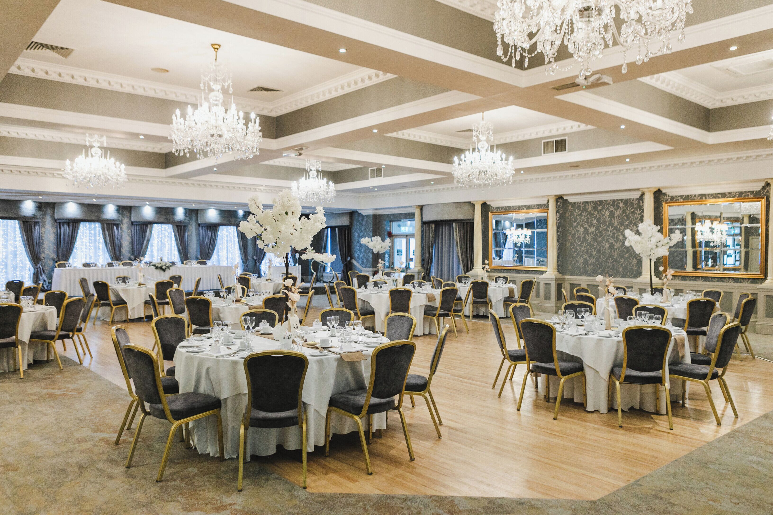 A spacious banquet hall with multiple round tables set for dining, elegant chandeliers, and large windows with curtains.