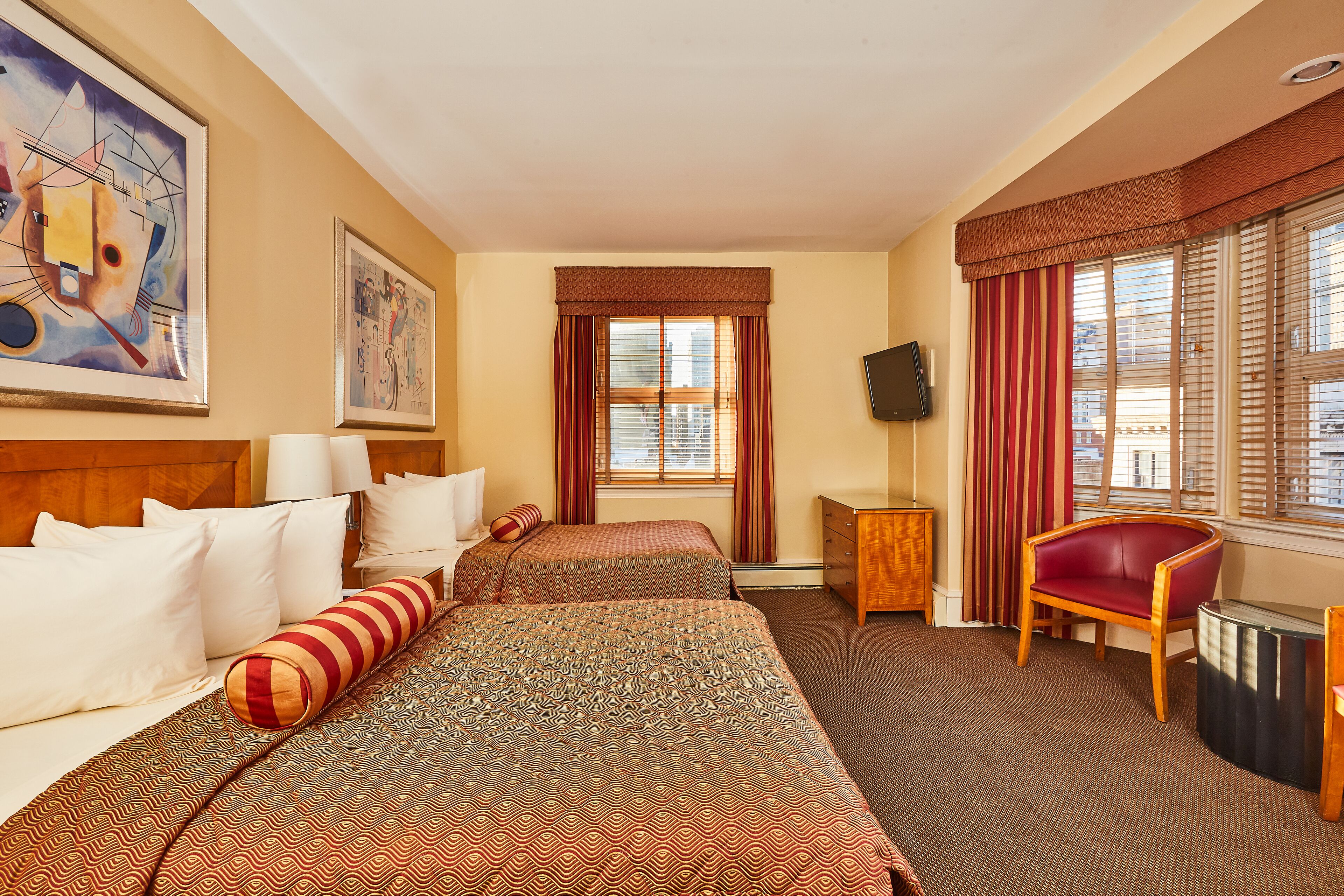 Habitación de hotel con dos camas, un escritorio, una silla, un televisor y obras de arte en las paredes.
