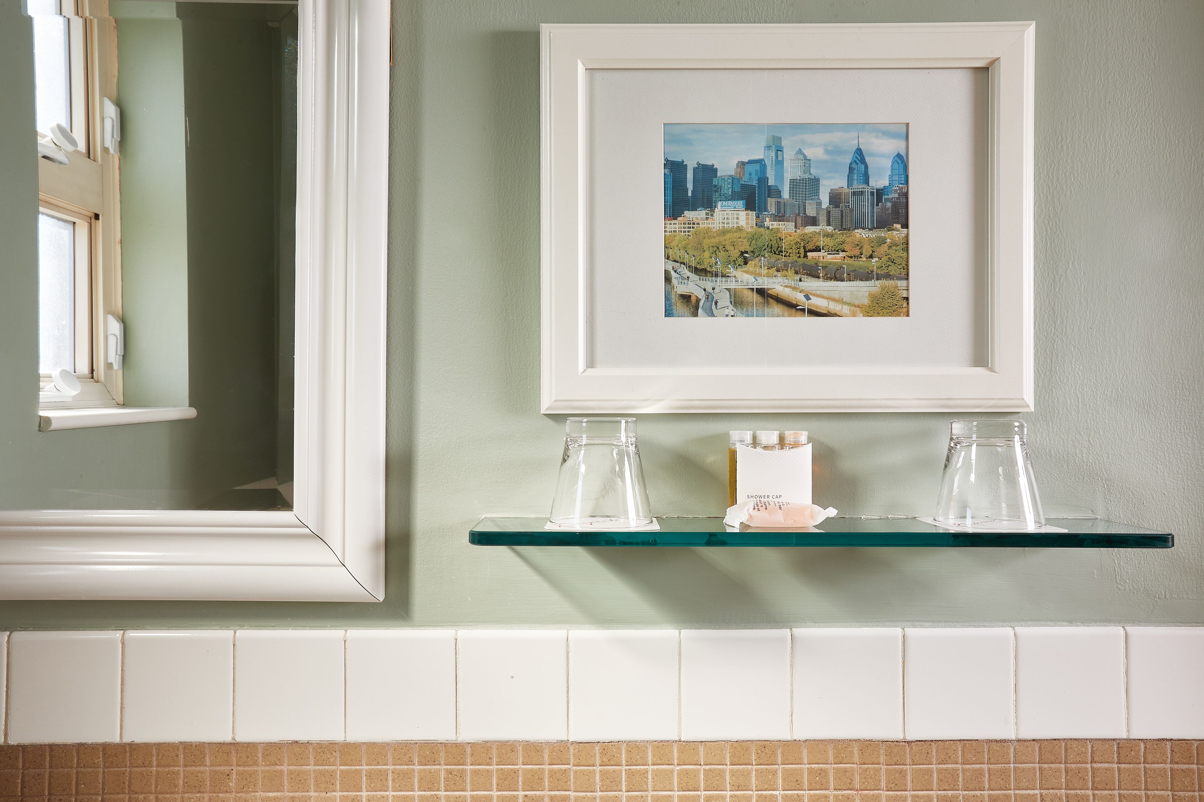 A framed cityscape photograph on a bathroom wall, a glass shelf with toiletries, and a mirror reflecting the scene.
