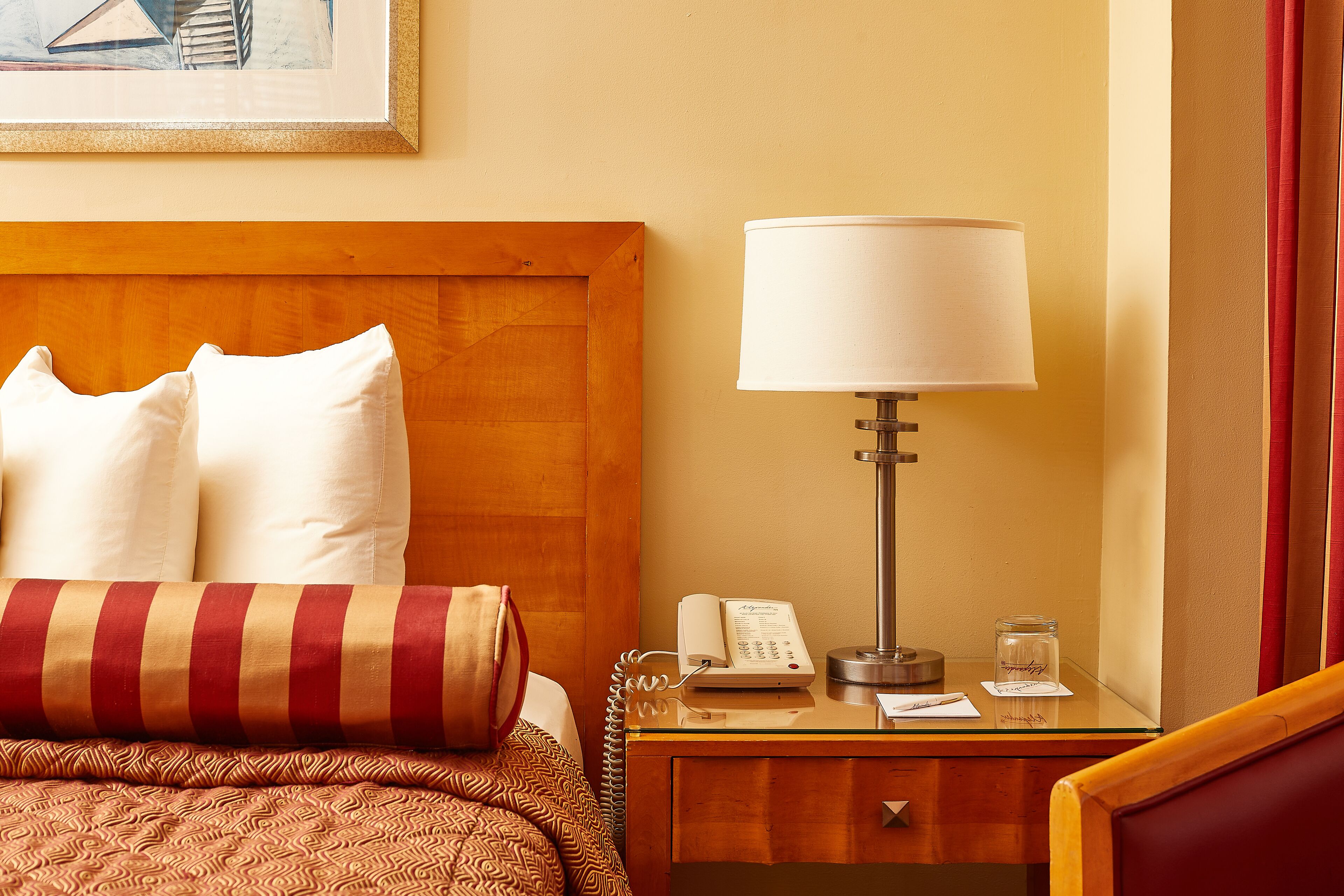 A neatly made bed with pillows and a striped blanket, a bedside lamp, a phone, and a small glass jar on a wooden nightstand.
