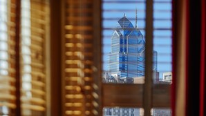Une vue à travers les stores d'une fenêtre, donnant sur un bâtiment moderne à la façade en verre et surmonté d'une flèche.