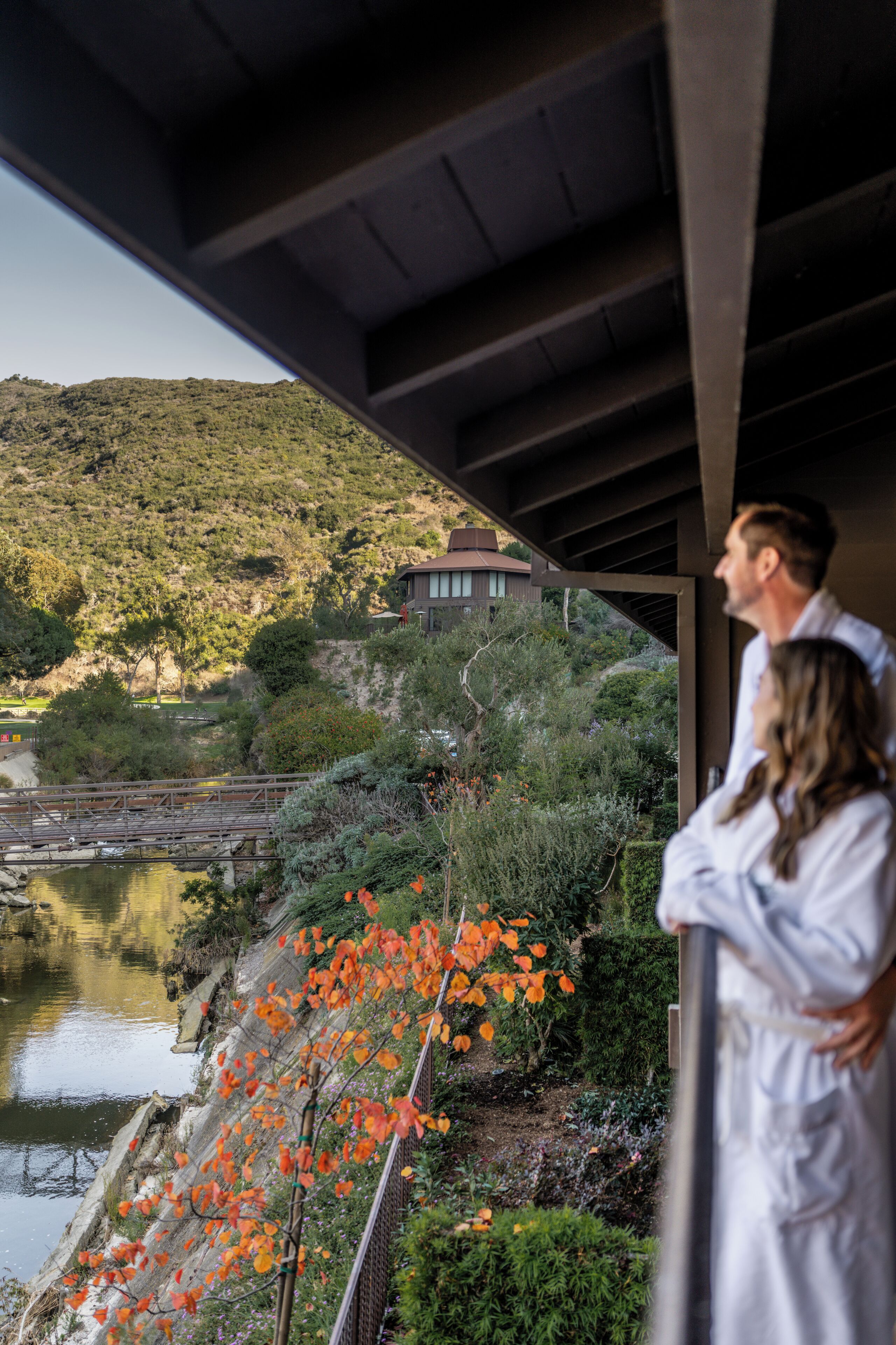 A view from a covered balcony overlooking a river, with a building on a hillside in the background.