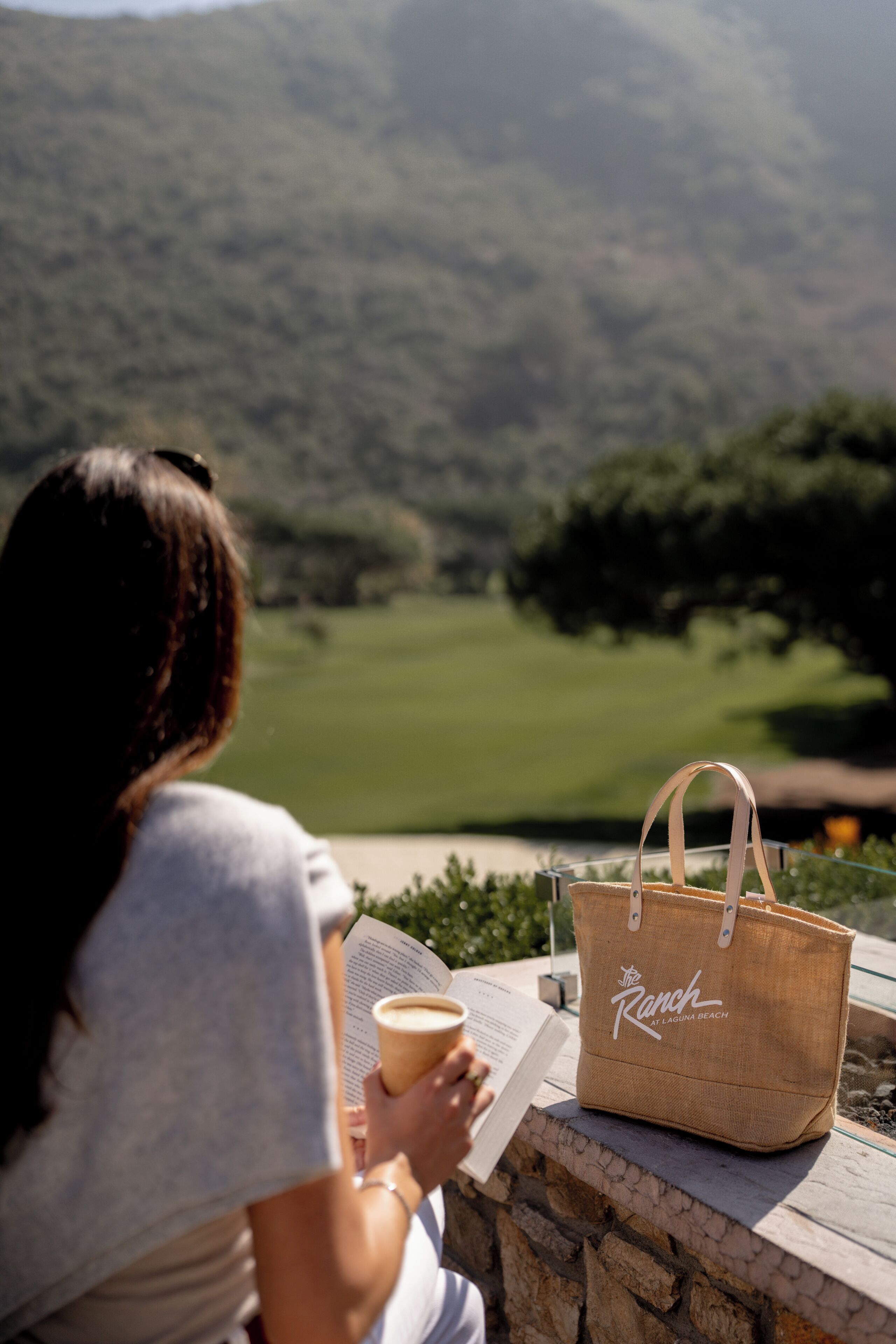 A person reading a book with a coffee cup, a straw bag, and a view of a green landscape.