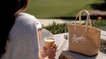 A person reading a book with a coffee cup, a straw bag, and a view of a green landscape.