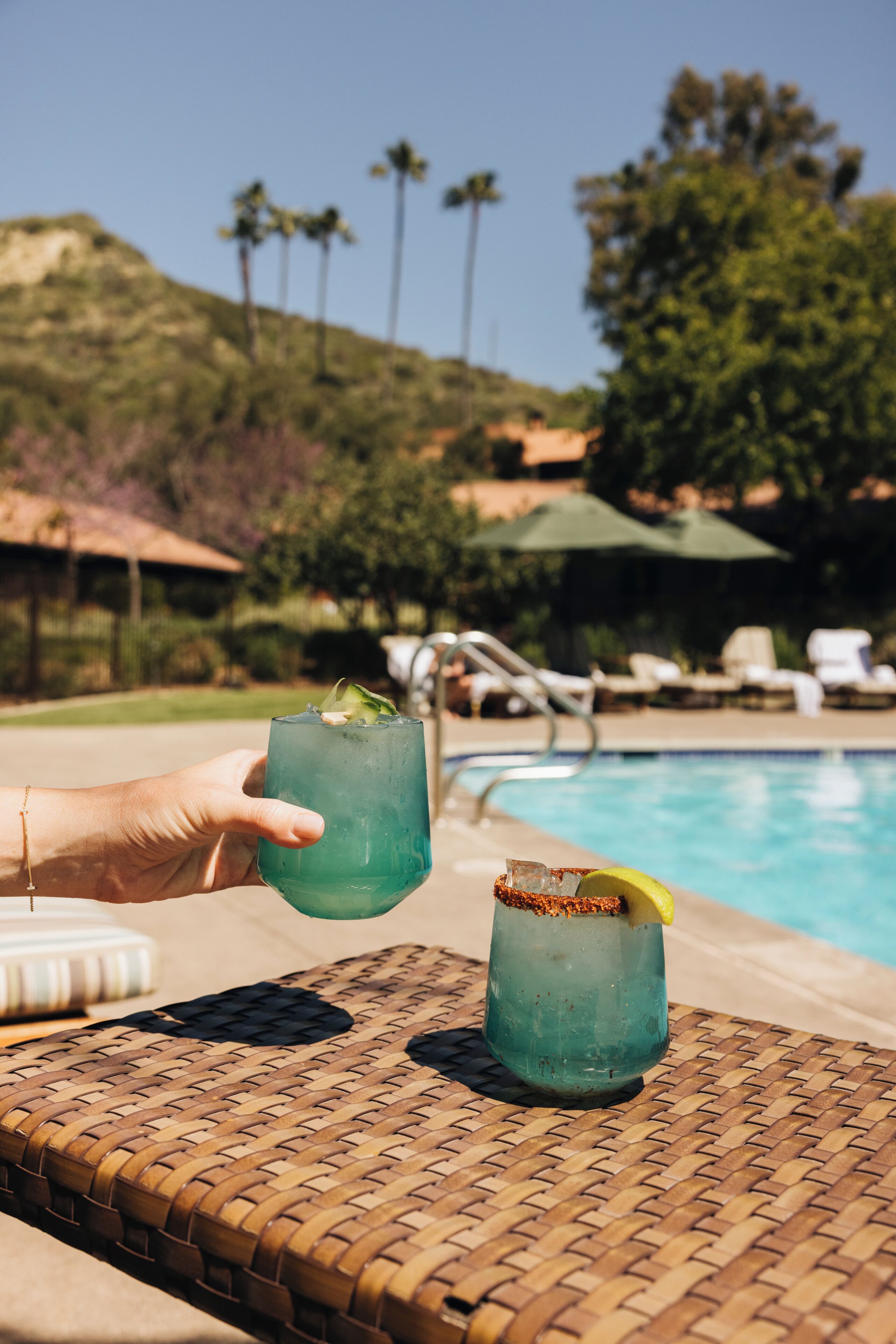 A poolside setting with a wicker table, two drinks, and a pool in the background.