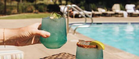 A poolside setting with a wicker table, two drinks, and a pool in the background.