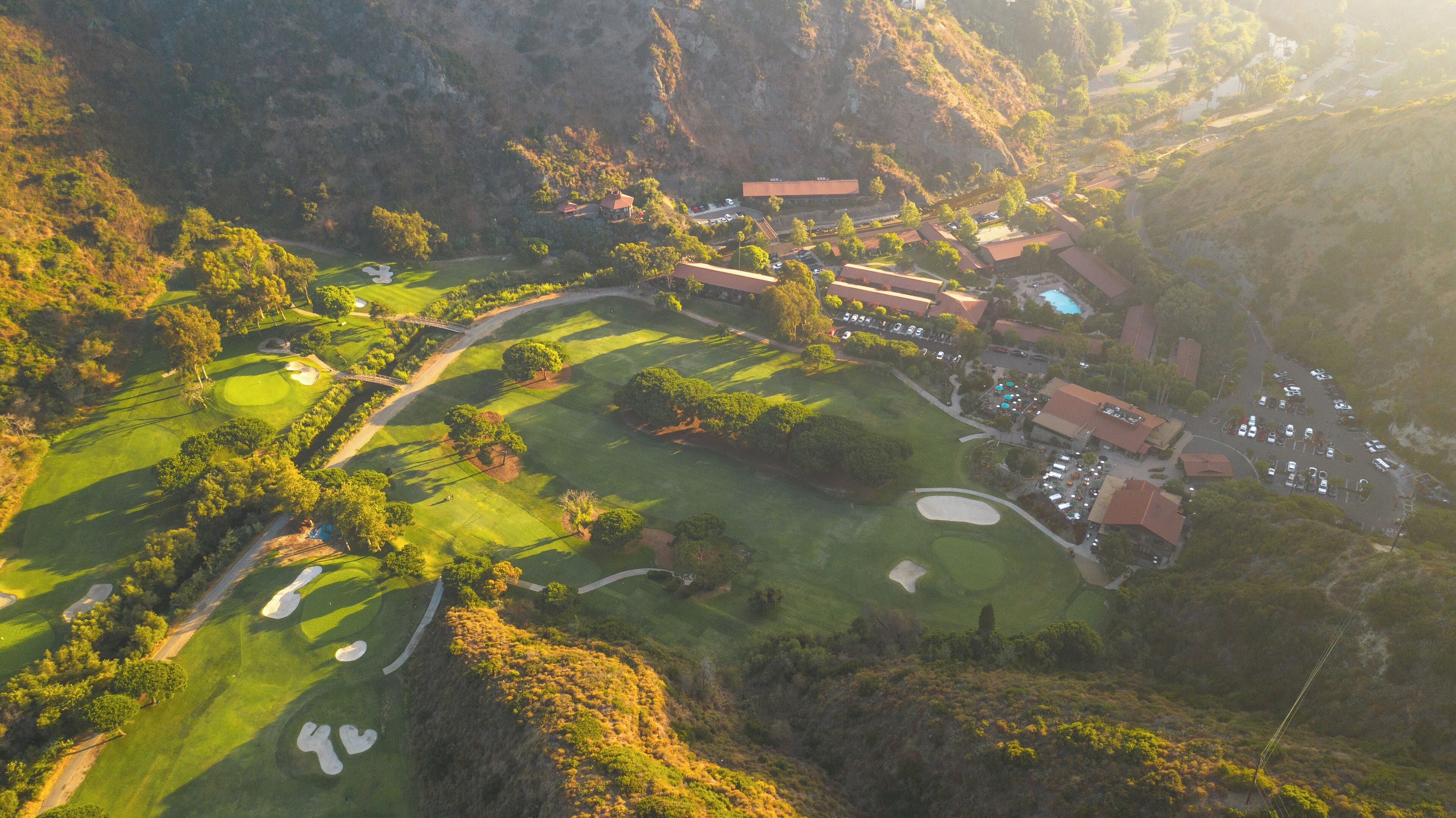 An aerial view of a golf course with a clubhouse, parking area, and surrounding landscape.