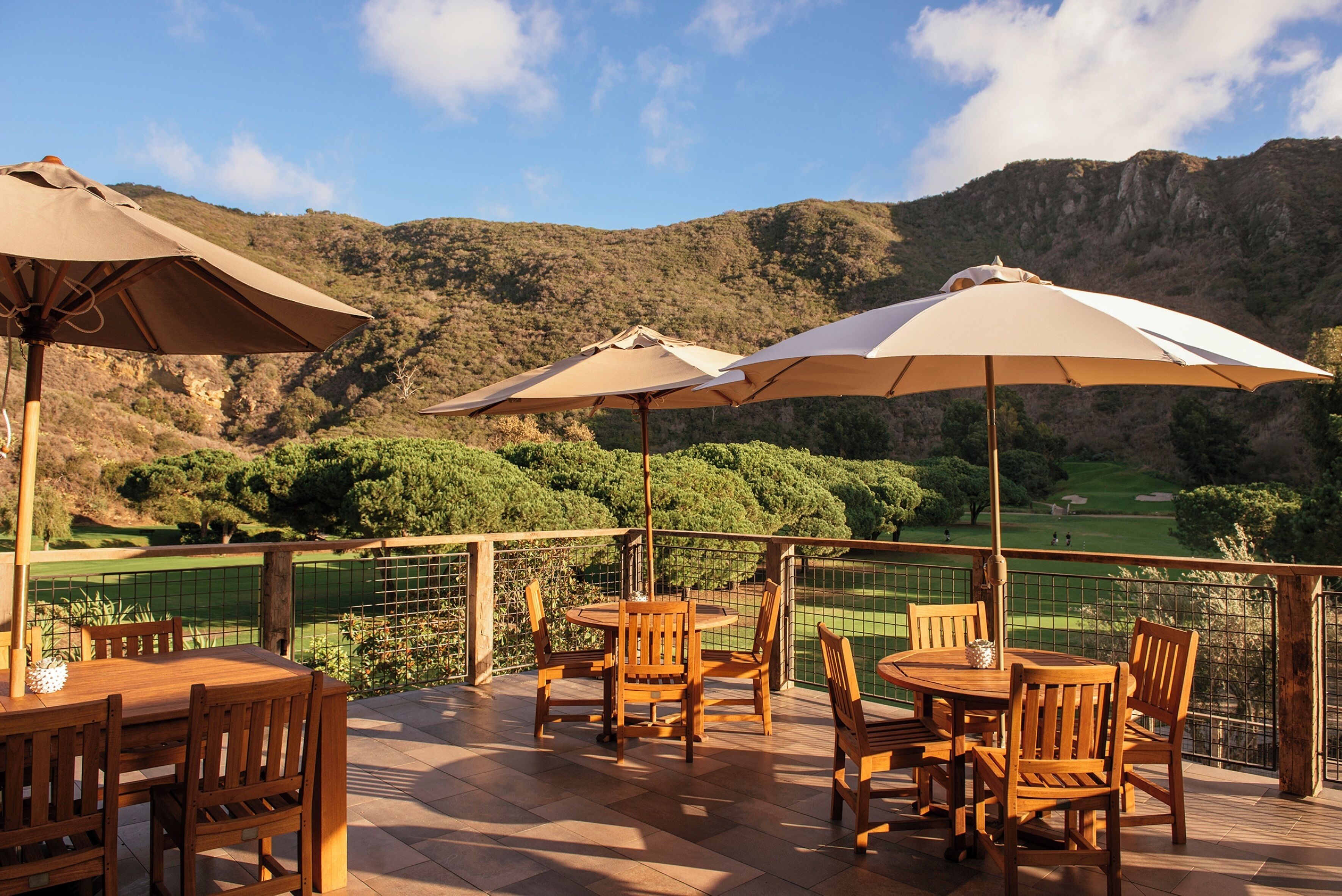 An outdoor patio with wooden tables and chairs, beige umbrellas, and a view of a golf course and mountains.