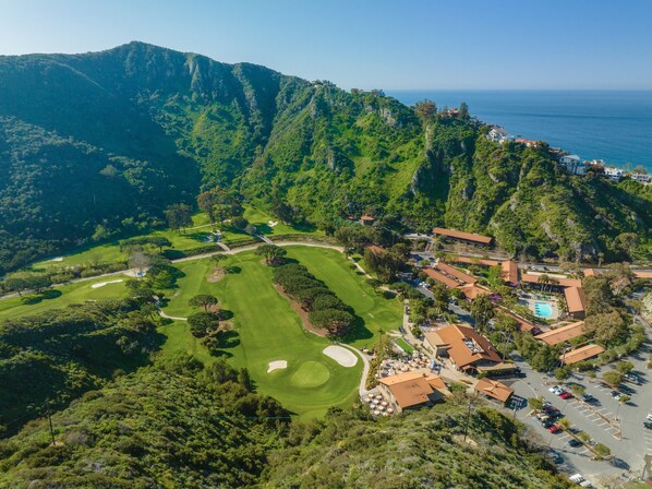 An aerial view of a golf course with a mountainous backdrop, a parking area, and buildings with red roofs. - The Ranch at Laguna Beach (Laguna Beach)