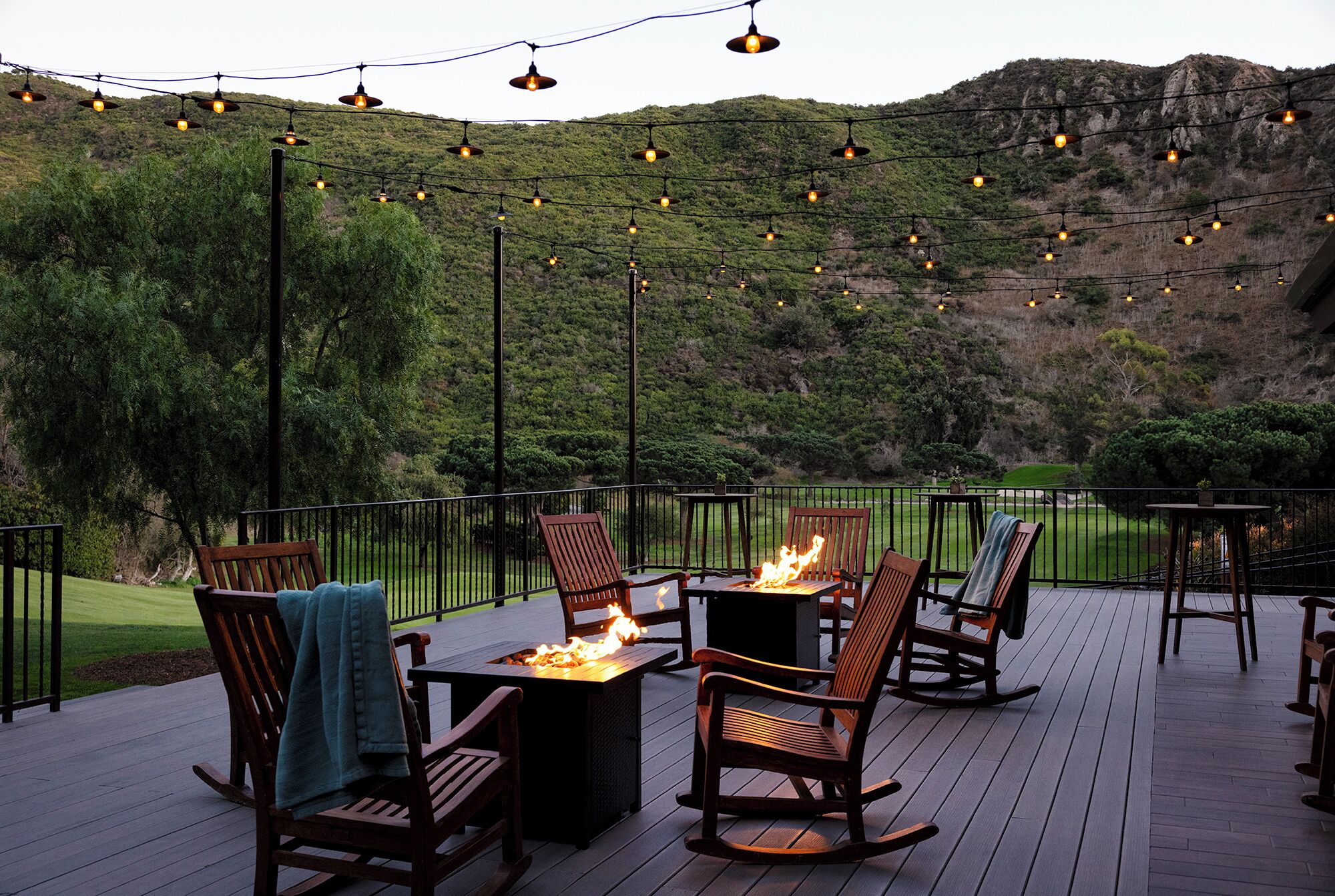 An outdoor deck with wooden furniture, a fire pit, and string lights against a backdrop of a hilly landscape.
