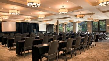 Un salón de conferencias con mesas y sillas dispuestas en filas, ventanas amplias e iluminación en el techo.