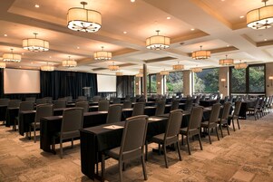 Un salón de conferencias con mesas y sillas dispuestas en filas, ventanas amplias e iluminación en el techo.