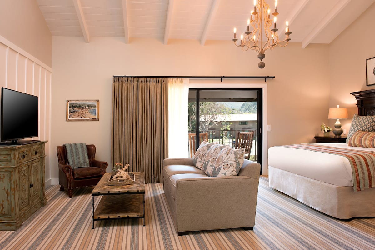 A hotel room with a large bed, a sofa, a television, a chandelier, and a view of the outdoors.