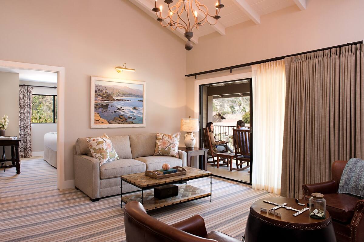 A living room with a sofa, coffee table, and chairs. There is a painting on the wall, a chandelier, and a view through the window.