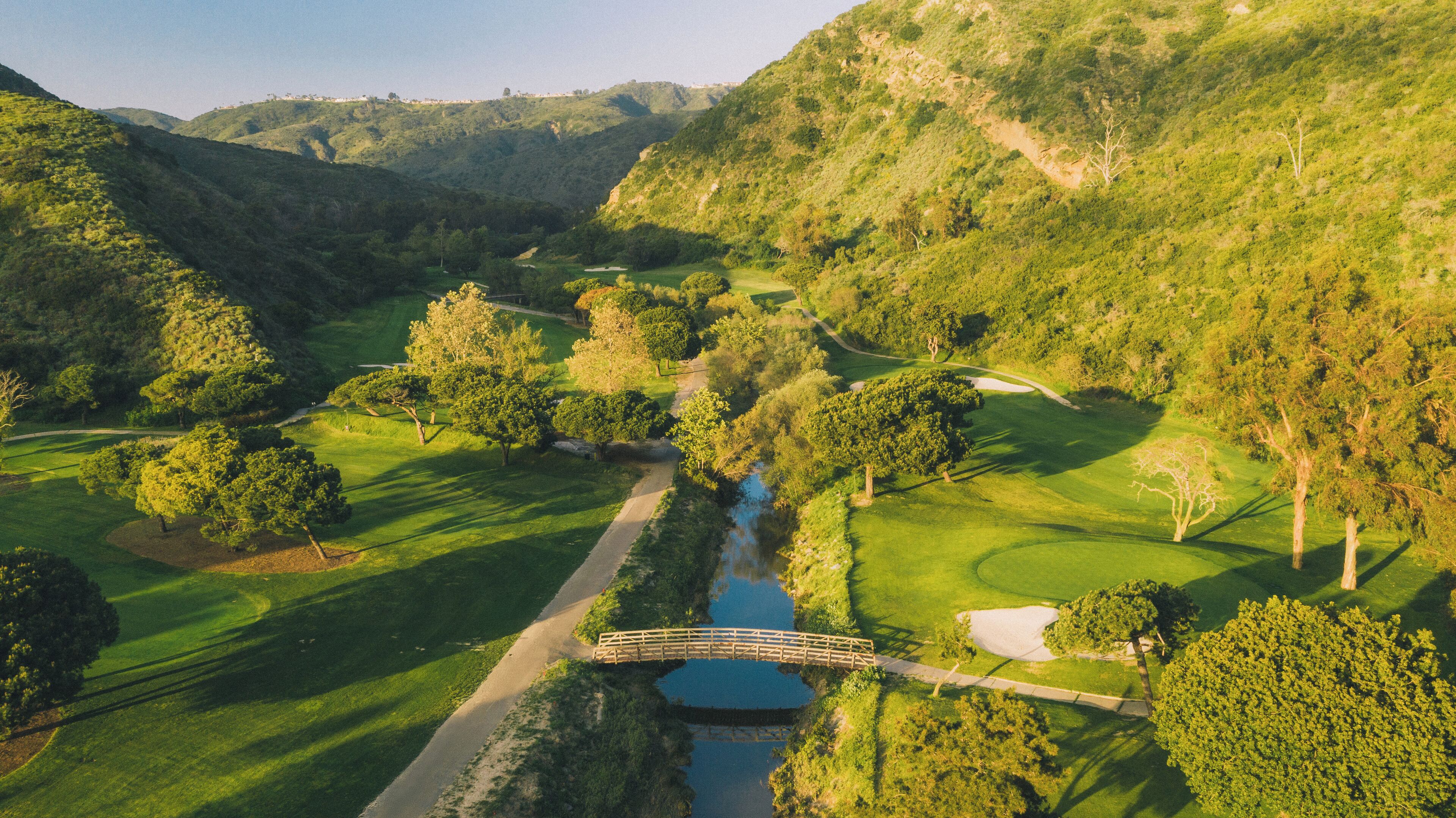 A golf course with a bridge over a stream, surrounded by lush greenery and hills.