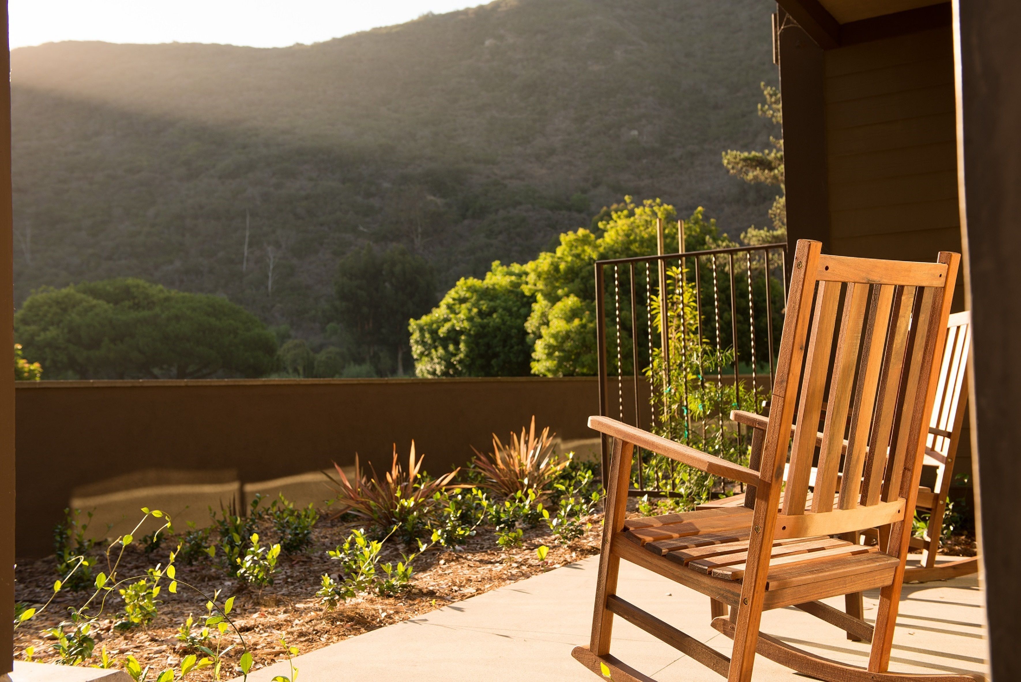 A wooden rocking chair on a balcony overlooking a mountainous landscape with a garden in the foreground.