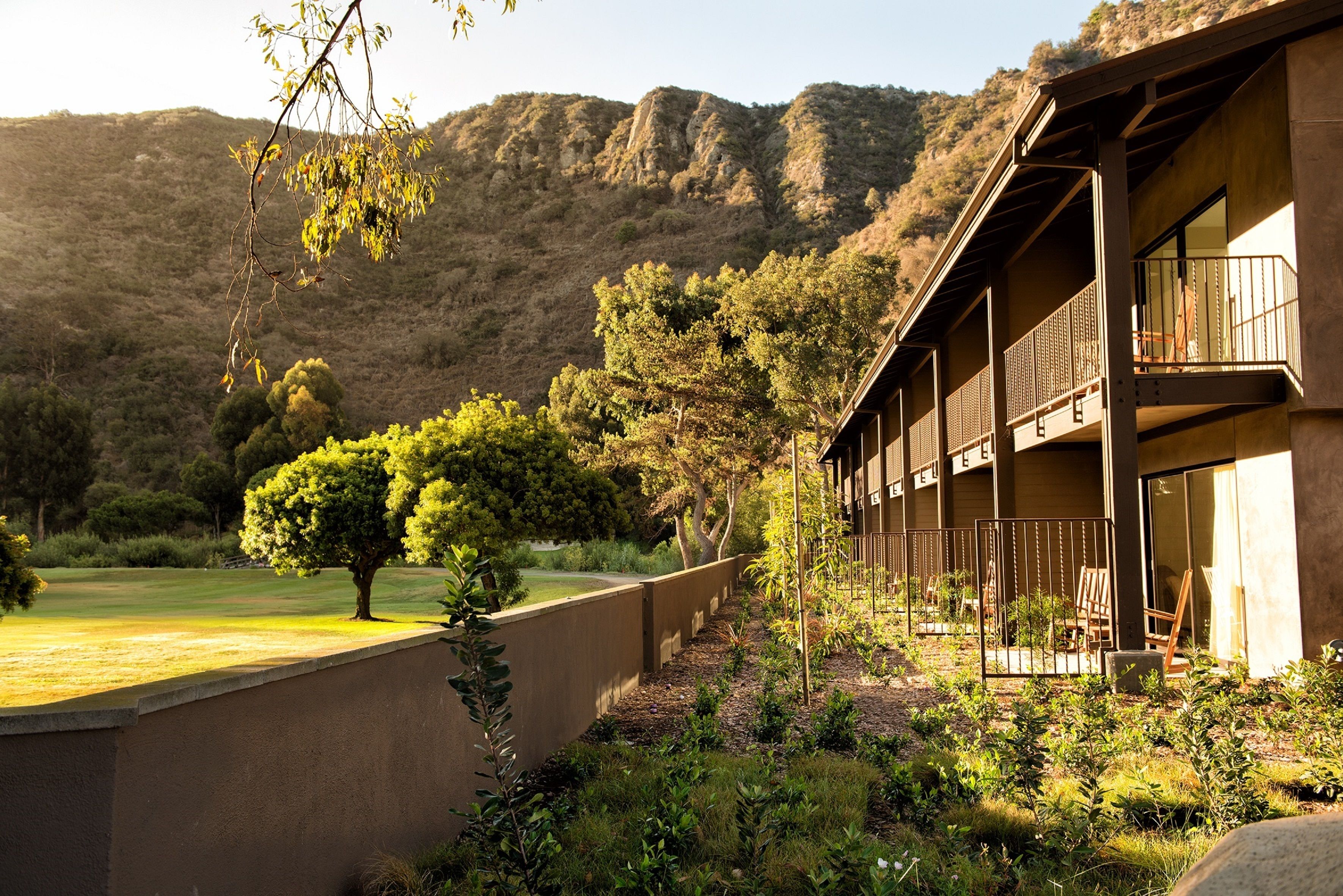 A building with a balcony, surrounded by a garden with trees and a view of a hilly landscape.