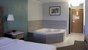 Quarto de hotel com uma banheira grande, uma cama, uma mesa de cabeceira com abajur e um quadro na parede.