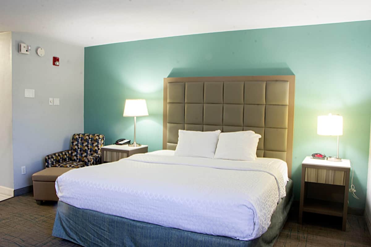 Une chambre d’hôtel avec un grand lit, une chaise, deux tables de chevet avec des lampes, une petite table et une horloge fixée au mur.