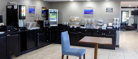 A modern hotel kitchen with a self-service breakfast area, including a refrigerator, microwave, and coffee station.