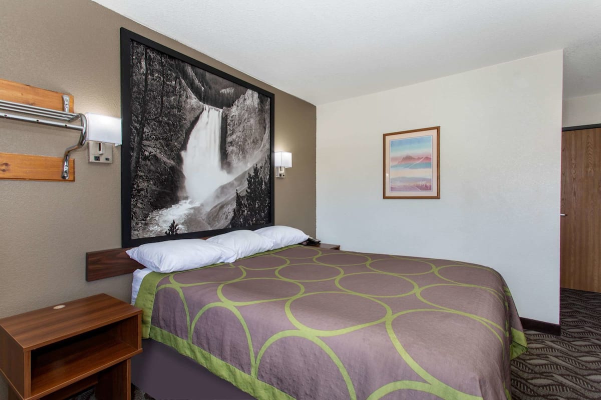 Ein Hotelzimmer mit einem großen Bett, einem hölzernen Kopfteil, einem Nachttisch, einer Wandleuchte, einem gerahmten Bild von einem Wasserfall und einem Teppichboden.