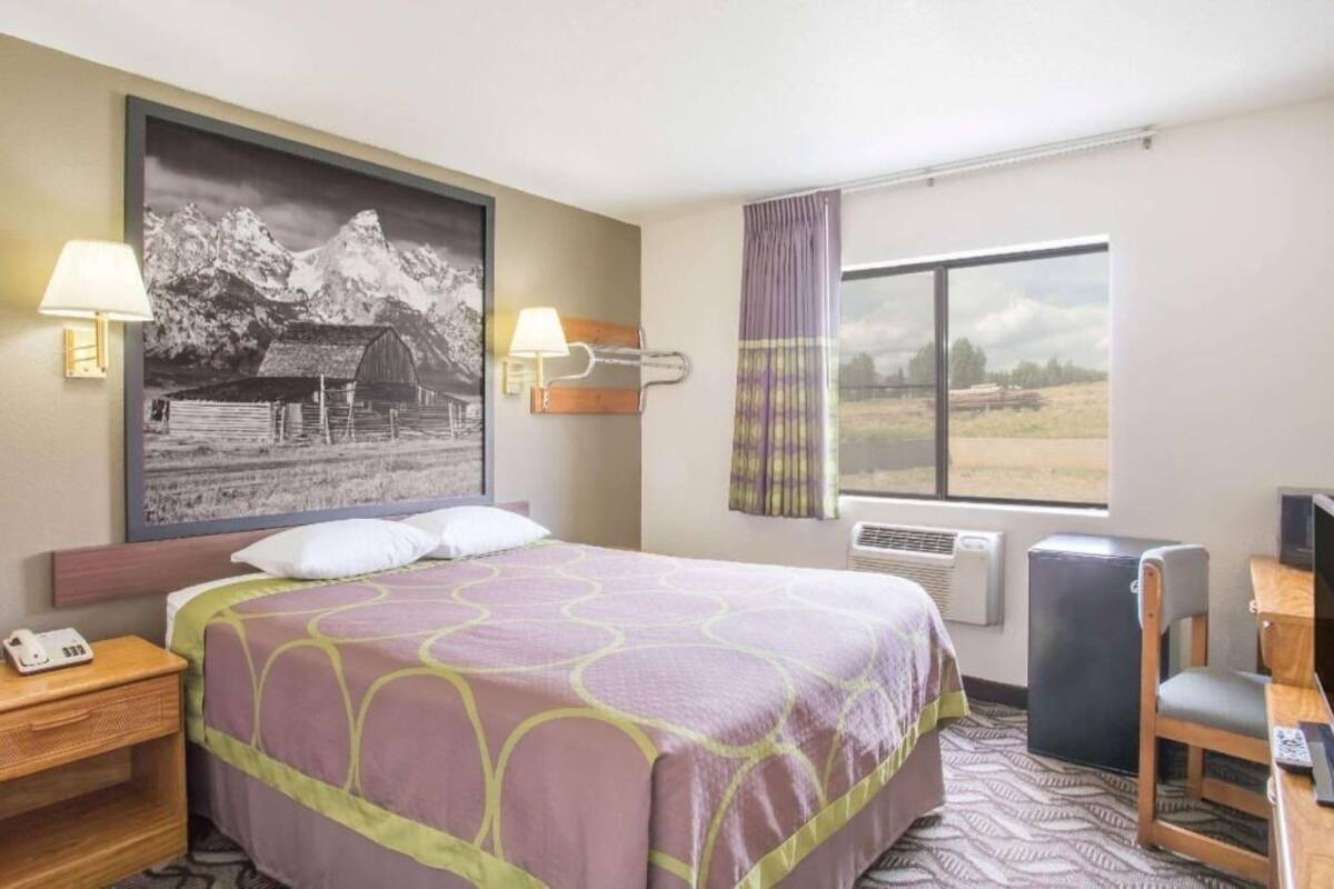 Ein Hotelzimmer mit einem Bett, zwei Nachttischlampen, einem großen gerahmten Bild, einem Fenster mit Vorhängen, einem Schreibtisch mit Stuhl und einem Fernseher.