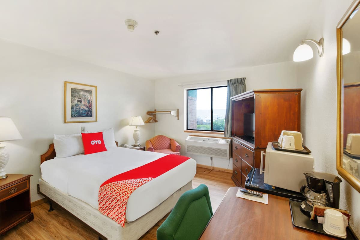 A hotel room with a large bed, a desk with a coffee maker, a chair, a TV, and a window with curtains.