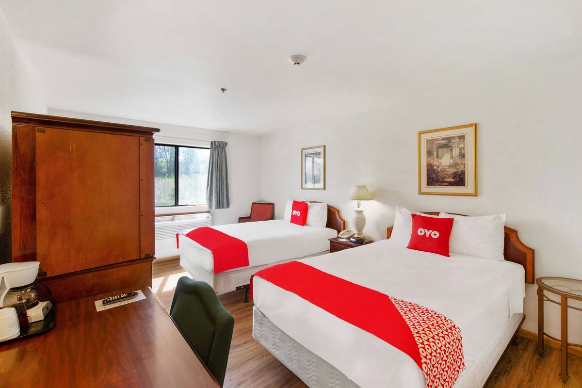 A hotel room with two beds, a wooden wardrobe, a desk with a coffee machine, and a window with curtains.
