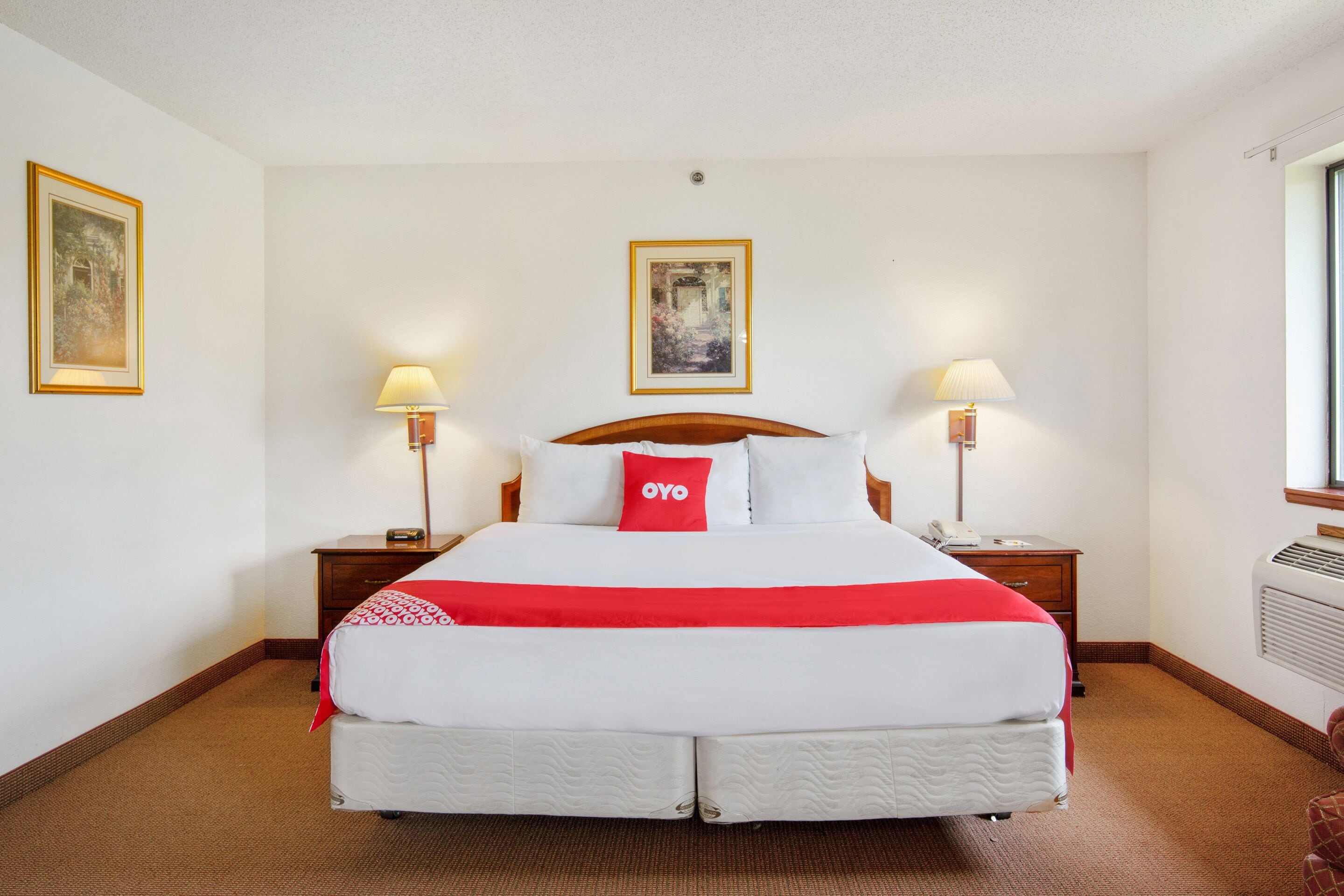 Quarto de hotel com uma cama grande, duas mesas de cabeceira, um criado-mudo, um ar condicionado fixado na parede e duas imagens emolduradas nas paredes.