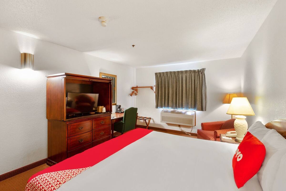A hotel room with a bed, a desk with a computer, a television, a window with curtains, a lamp, and a red throw blanket.