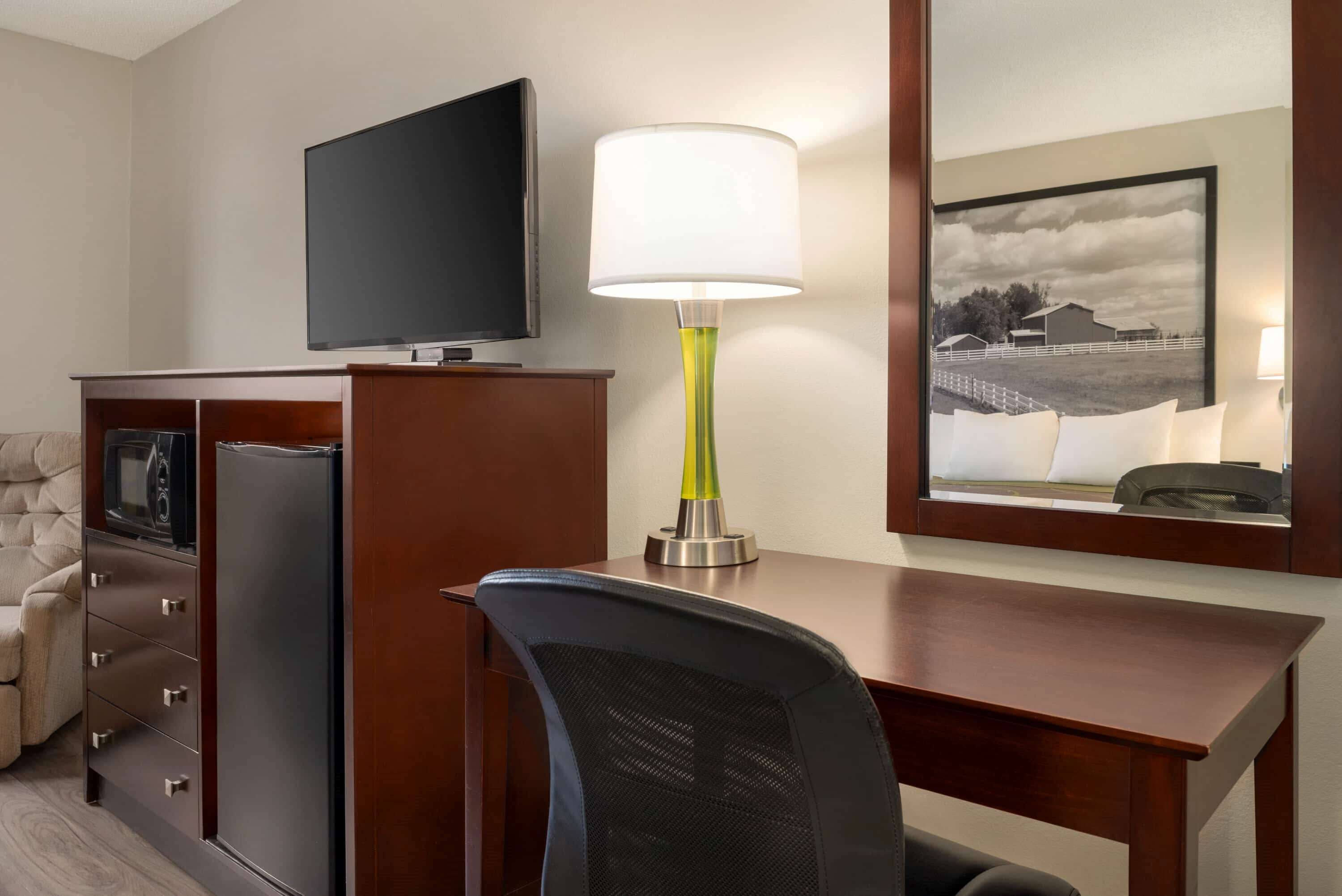 A hotel room with a wooden desk, a television, a lamp, a chair, and a framed picture on the wall.