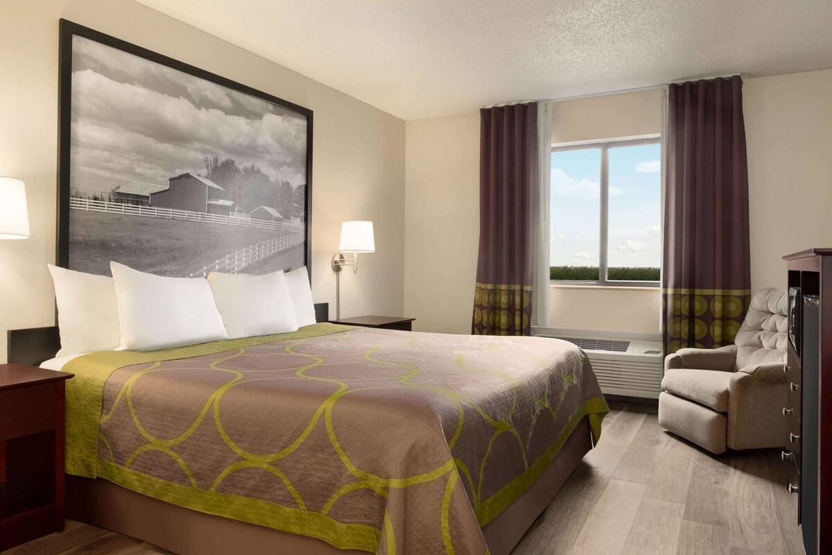 Une chambre d’hôtel avec un grand lit, une chaise, un bureau, une grande fenêtre avec des rideaux, une lampe fixée au mur et un grand tableau encadré.