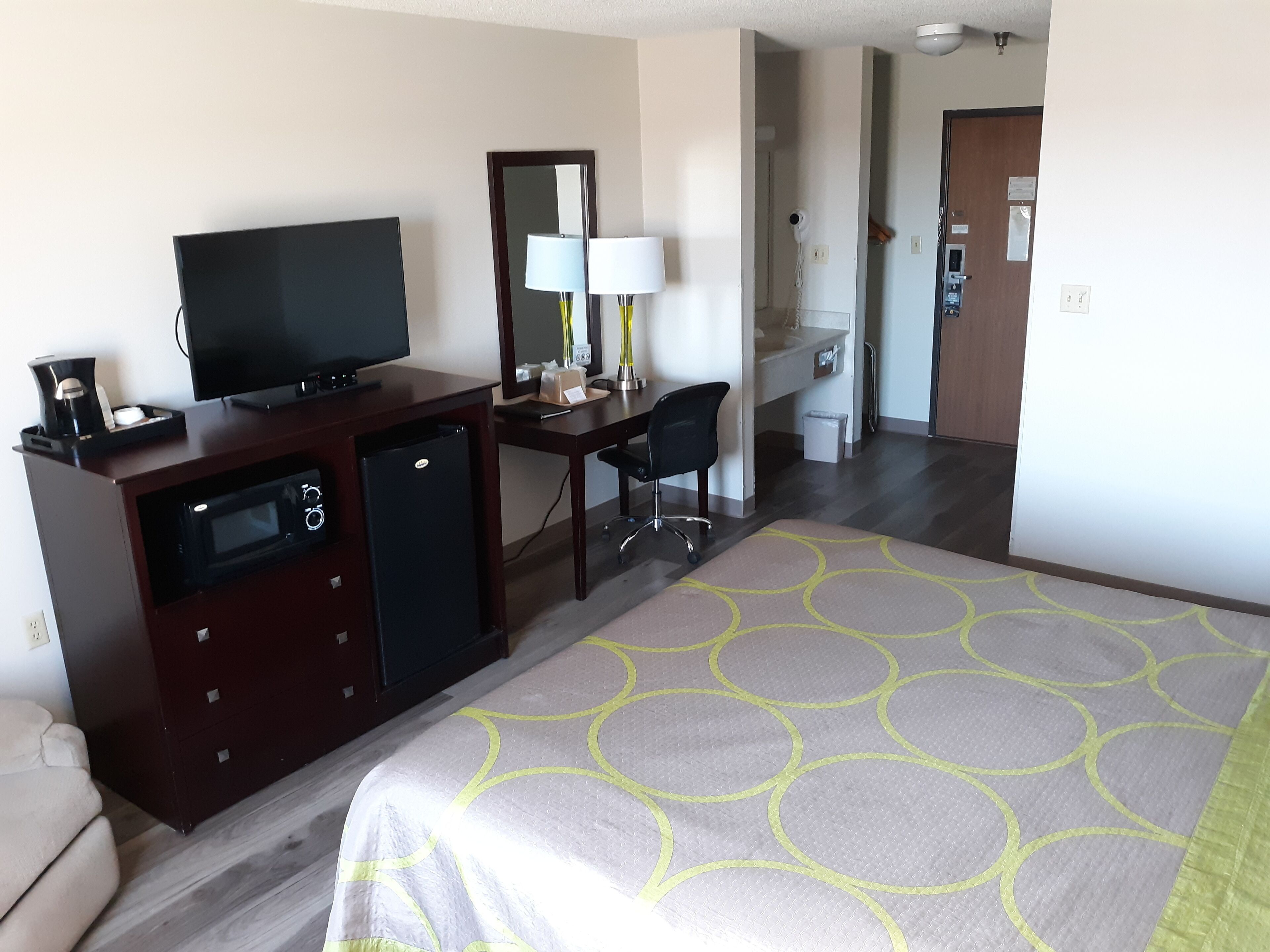 A hotel room with a bed, a desk with a chair, a television, a mini-fridge, and a bathroom visible through an open door.