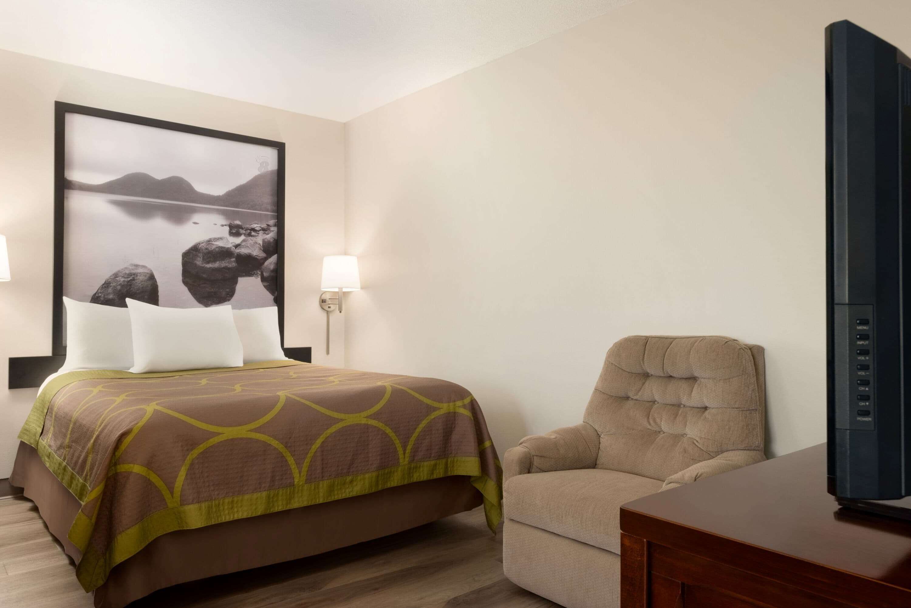 A hotel room with a bed, a chair, a television, and a wall-mounted lamp.